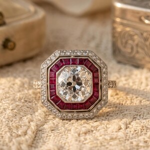 May include: An octagonal ring featuring a central round faceted diamond surrounded by a border of square-cut red rubies. The outer edge of the ring is framed by a halo of small round diamonds set in a metallic band with intricate milgrain detailing.