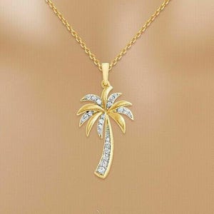 May include: Gold-toned necklace featuring a palm tree pendant. The pendant is adorned with sparkling white gemstones, and the chain is a delicate gold link. The palm tree design is a symbol of tropical vacations and summer.