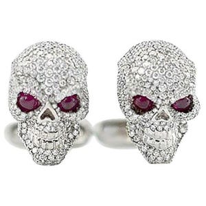 May include: Two silver skull rings encrusted with clear gemstones. The eye sockets are filled with deep red gemstones. The rings are set against a white background.