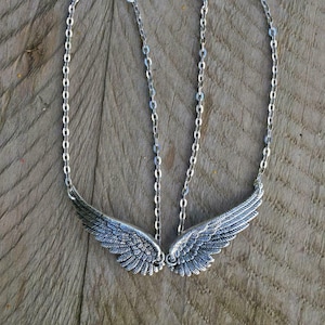 Op de afbeelding: Een zilverkleurige ketting met een paar gedetailleerde vleugelhangers. De vleugels zijn verbonden door fijne kettingen en sluitingen. De ketting wordt gepresenteerd op een rustiek houten oppervlak, met de gedetailleerde veren.