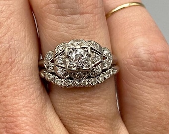 Könnte beinhalten: Ein Vintage-Verlobungsring mit Diamanten, einem zentralen Stein und einem zarten Band. Der Ring ist in Weißgold gefasst und hat ein florales Design.