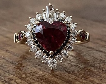 Könnte beinhalten: Ein goldener Ring mit einem herzförmigen, tiefroten Edelstein im Zentrum. Der Edelstein ist von einem Kranz aus weißen und schwarzen Steinen umgeben, mit zusätzlichen roten Steinen an den Schultern des Rings. Auf Holzgrund.