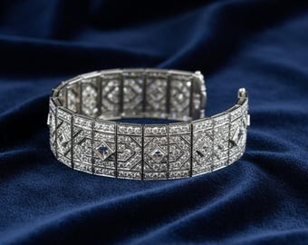 May include: A silver bracelet with a geometric design, encrusted with numerous small, sparkling gemstones. The bracelet is open, showcasing its intricate pattern against a deep blue velvet backdrop. The gemstones reflect light, creating a dazzling effect.