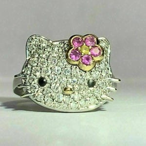 May include: A silver ring with a Hello Kitty design. The ring is covered in small diamonds and features a pink flower with yellow center.