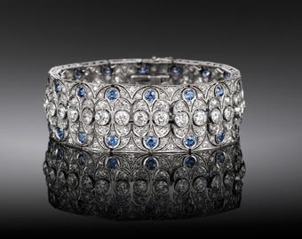 May include: A wide, ornate bracelet with a silver metal setting. The bracelet is encrusted with numerous clear, round-cut gemstones and interspersed with small, round blue gemstones. The bracelet is displayed on a reflective surface.