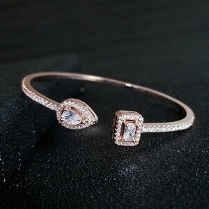 Könnte beinhalten: Offener Armreif aus Roségold mit birnenförmigem und quadratischem, klarem Steinfassungen. Das Armband ist mit kleinen, klaren Steinen besetzt. Der Schmuck wird auf einer schwarzen Oberfläche präsentiert.