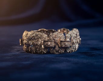 May include: An ornate silver bracelet with intricate floral and geometric designs. The bracelet features multiple rectangular and circular links, each adorned with small, sparkling gemstones. The piece is set against a dark blue velvet background, highlighting its elegant details.