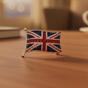 May include: A silver-toned brooch in the shape of the Union Jack flag. The flag design features red and blue gemstones, with a silver pole. The brooch is set on a wooden surface.