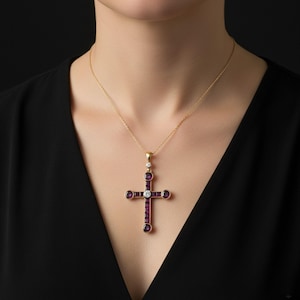 May include: A gold-toned cross pendant necklace. The cross features a central row of rectangular, deep red gemstones, with round red gemstones at the ends of each arm. A single white gemstone is set in the center, and a white gemstone and red gemstone are set above the cross.