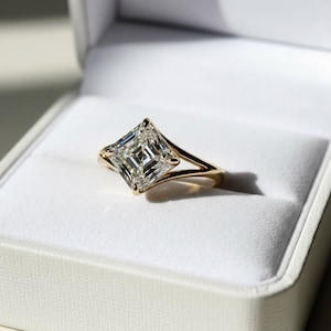 May include: A gold engagement ring with a large, square-cut diamond. The ring is set in a gold band with a split shank design. The ring is displayed in a white ring box with a soft, velvety interior.