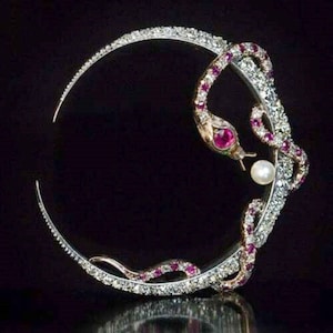 Puede incluir: Broche en forma de media luna con diseño de serpiente. El cuerpo de la serpiente está adornado con pequeños diamantes y rubíes brillantes. La cabeza de la serpiente tiene un ojo de rubí y una perla. La broche está sobre fondo negro.