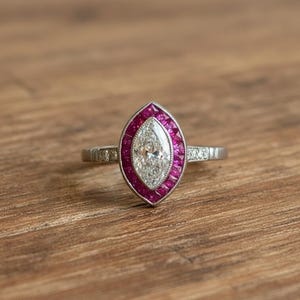 May include: A silver-toned ring featuring a central marquise-cut clear gemstone surrounded by a halo of small, rectangular-cut vibrant pink rubies. The metal band includes small, round, clear accent stones set along the sides of the shank.