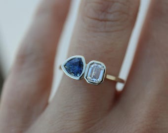 May include: A gold ring featuring two gemstones: a triangular blue sapphire and a rectangular, clear emerald-cut diamond. The gemstones are set side-by-side in a bezel setting. The ring is shown on a person's finger.