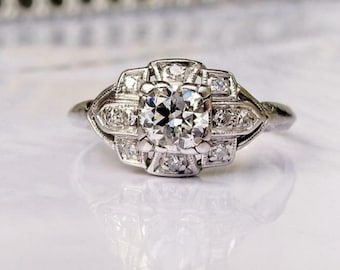 May include: A close-up of a silver ring with a large, round, clear diamond center stone. The ring has a geometric design with smaller diamonds surrounding the center stone. The ring is set on a white surface.