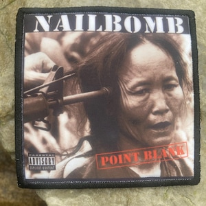 May include: Square patch featuring a sepia image of a person with a gun aimed at their head. The text "NAILBOMB" is at the top, and "POINT BLANK" is in a red stamp. The patch has a black border and an advisory warning.