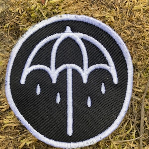 May include: A circular black embroidered patch featuring a white umbrella design with three raindrops. The patch has a white border and is likely made of fabric.