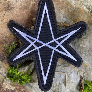 May include: Black embroidered patch with a white geometric design. The patch has a star-like shape with a central diamond and intersecting lines. The edges are neatly finished with black thread, suitable for clothing or accessories.