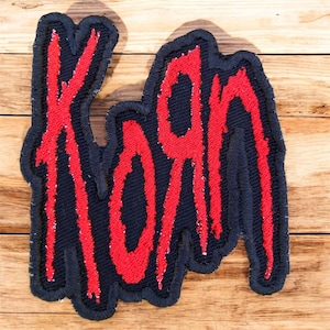 May include: Embroidered patch with the word "KORN" in a dripping red font. The text is on a navy blue background with a matching border. This patch is designed for application on clothing or accessories.