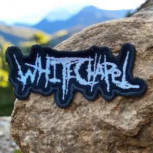May include: A black embroidered patch featuring the band name "WHITECHAPEL" in light blue, stylized lettering. The patch has a dark blue border and is set against a natural, rocky background with a blurred mountain view.