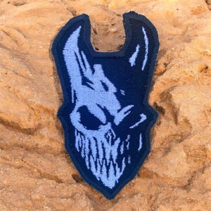 May include: A dark blue and light blue embroidered patch with a skull design. The patch has a light blue skull graphic with sharp teeth and a menacing expression, set against a dark blue background. The patch is on a textured, tan surface.