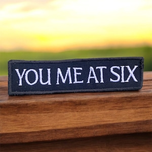 May include: A dark blue rectangular patch with the white embroidered text "YOU ME AT SIX". The patch is placed on a wooden surface, with a blurred green and yellow background.