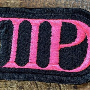 May include: Embroidered patch featuring the letters "IIP" in bright pink, on a black background. The patch has a black border and is likely designed to be sewn onto clothing or accessories. The design is striking and would stand out.