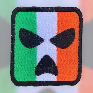 May include: Embroidered patch depicting the Irish flag with a black, stylized face. The patch has a green, white, and orange striped background, with a black border. The face features two large, angled eyes and a downturned mouth.
