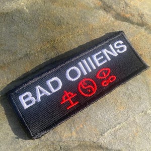 May include: Black rectangular patch with the words "BAD OMENS" embroidered in white. Red symbols are below the text. The patch is on a textured grey stone surface. The patch is likely for clothing or accessories.