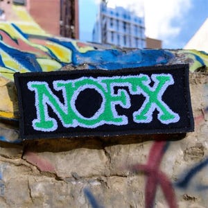 May include: A black rectangular patch featuring the word "NOFX" in green and white embroidery. The patch is set against a weathered stone wall with graffiti. The patch is a band logo, suitable for clothing or accessories.