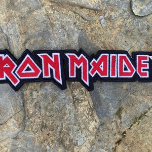 May include: A rectangular embroidered patch displaying the words "IRON MAIDEN" in red capital letters with a white border, on a black backing. The patch is designed for application to clothing or accessories.