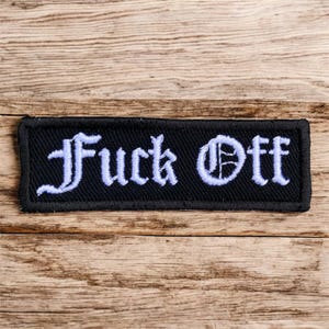 May include: A black rectangular patch with the words "Fuck Off" embroidered in white, in a gothic font. The patch has a black border and is set against a wooden background.