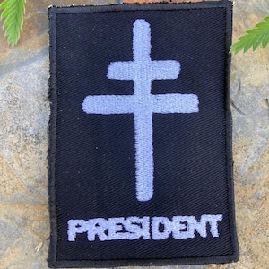 May include: Black rectangular patch featuring a light blue double cross and the word "PRESIDENT" in white. The patch has a black border. It is likely an embroidered or woven item.