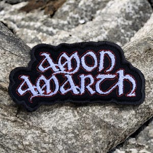 May include: A black embroidered patch featuring the band name "AMON AMARTH" in a stylized font. The lettering is light blue with a red outline, and the patch has a black border. The patch is displayed on a gray, textured rock surface.