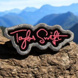 May include: Embroidered patch featuring the name "Taylor Swift" in pink script on a black base, edged with a grey border. The patch is shown on a rock, with a mountain range in the background.