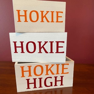 May include: Three white wooden boxes stacked on top of each other. The top box says "Hokie" in orange letters. The middle box says "Hokie" in red letters. The bottom box says "Hokie High" in orange and red letters.