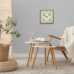 May include: A white clock with black hands and Roman numerals on a beige background with a green and gold border. The clock is hanging on a gray wall above a white coffee table with a white teapot and two white cups. A white throw blanket is draped over a wooden chair with a white cushion.