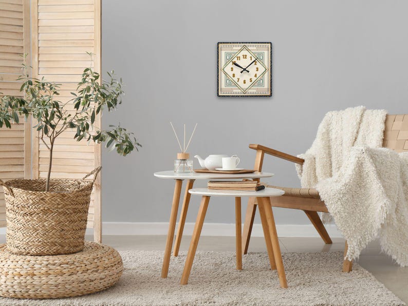 May include: A white clock with black hands and Roman numerals on a beige background with a green and gold border. The clock is hanging on a gray wall above a white coffee table with a white teapot and two white cups. A white throw blanket is draped over a wooden chair with a white cushion.