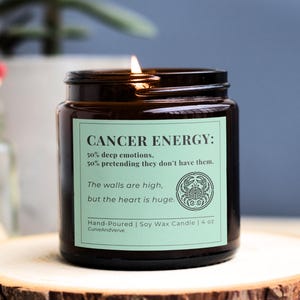 Cancer Zodiac Candle with light green label, displaying humorous Cancer energy breakdown. Hand-poured soy wax candle in amber glass jar, lit with a cozy flame on wooden stand.