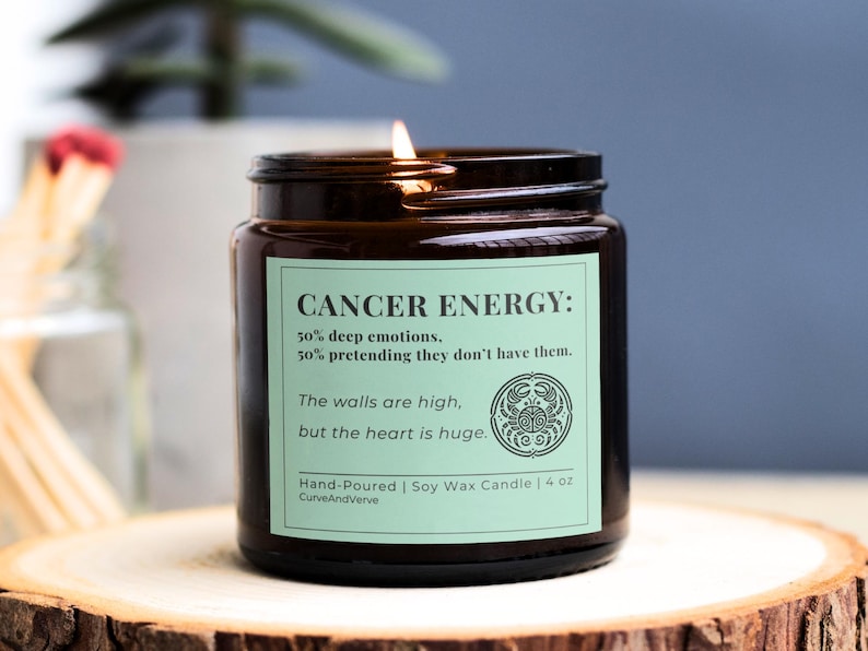 Cancer Zodiac Candle with light green label, displaying humorous Cancer energy breakdown. Hand-poured soy wax candle in amber glass jar, lit with a cozy flame on wooden stand.