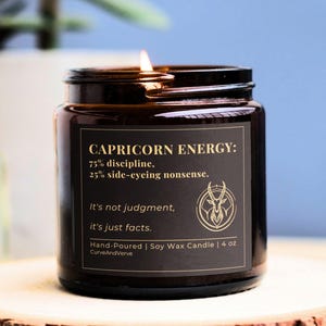 Capricorn Energy Candle with gold lettering on a dark background, burning softly on a wooden coaster, showcasing the label with the phrases "75% discipline, 25% side-eyeing nonsense" and "It's not judgment, it's just facts."