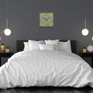 Sage green and gold Art Deco square clock displayed above a modern bed with crisp white linens, flanked by black nightstands, gold-accented planters, and pendant globe lights.