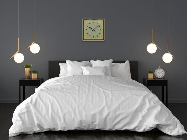 Sage green and gold Art Deco square clock displayed above a modern bed with crisp white linens, flanked by black nightstands, gold-accented planters, and pendant globe lights.