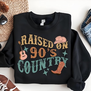 Può includere: Felpa nera con una grafica in stile vintage che dice "Raised on 90's Country". La grafica include una chitarra, un cappello da cowboy e uno stivale da cowboy.