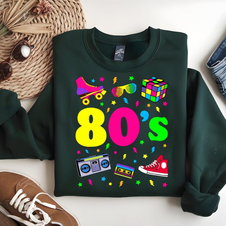 May include: A dark green sweatshirt with a colorful 80's themed graphic. The graphic includes a roller skate, sunglasses, a Rubik's cube, a boombox, a cassette tape, and a pair of sneakers. The text "80's" is in large, colorful letters.
