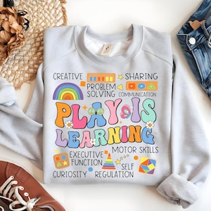 May include: A light gray sweatshirt with the text "Play is Learning" in colorful, retro-style letters. The text is surrounded by illustrations of a rainbow, a butterfly, flowers, and peace signs. Below the text, there are a series of colorful icons and words that describe the benefits of play, including "creative", "sharing", "problem solving", "communication", "executive function", "motor skills", "curiosity", and "self regulation."