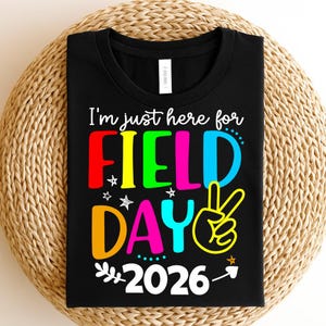 May include: Black t-shirt with the text "I'm just here for FIELD DAY 2026" in colorful letters. The design includes a peace sign and stars. The shirt is displayed on a woven, circular surface.