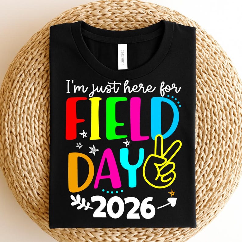May include: Black t-shirt with the text "I'm just here for FIELD DAY 2026" in colorful letters. The design includes a peace sign and stars. The shirt is displayed on a woven, circular surface.