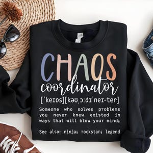 Może przedstawiać: Czarny sweter z napisem "CHAOS coordinator" w kolorowych literach, zdefiniowany jako "Ktoś, kto rozwiązuje problemy, o kt&oacute;rych nigdy nie wiedziałeś, że istnieją, w spos&oacute;b, kt&oacute;ry cię oszołomi; Zobacz także: ninja; rockstar; legenda".