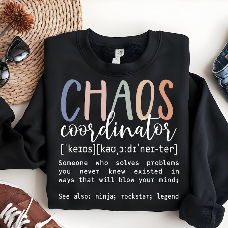 Może przedstawiać: Czarny sweter z napisem "CHAOS coordinator" w kolorowych literach, zdefiniowany jako "Ktoś, kto rozwiązuje problemy, o kt&oacute;rych nigdy nie wiedziałeś, że istnieją, w spos&oacute;b, kt&oacute;ry cię oszołomi; Zobacz także: ninja; rockstar; legenda".