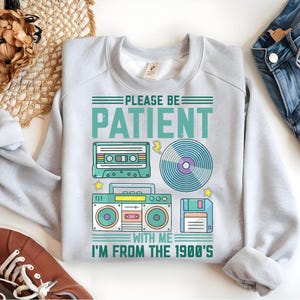 May include: Gray sweatshirt with a retro design featuring a cassette tape, record, boombox, and floppy disk. The text reads "Please be patient with me I'm from the 1990's".